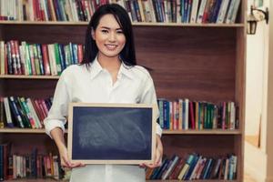 beautiful young woman holding the blackboard in front of bookshelf and smiling to camera photo