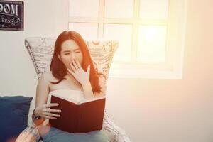 Beautiful young woman sitting on sofa and yawning near the window tired of reading photo