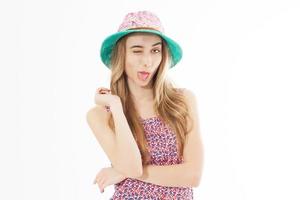 chica joven positiva con sombrero de verano de color posando sobre un fondo blanco y mostrando la lengua. foto