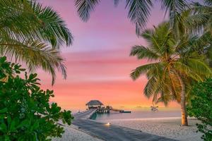 isla turística de maldivas al atardecer con embarcadero de madera, increíble cielo colorido. perfecto paisaje de playa al atardecer. detalle de hojas de palma en primer plano. vacaciones y relajación en la playa, fondo de vacaciones de verano foto