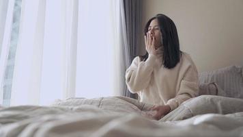 Young adult Asian woman wakes up yawning and Stretch her arm on the bed, lazy morning getting up late on the weekend day off, comfortable bedroom window curtains, natural day light, urban lifestyle video