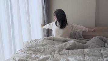 bred bild av ung vacker asiatisk kvinna sätter sig på sängen och sträcker ut armarna, kopplar av på helgmorgonen, bekväma sovrumsfönstergardiner, naturligt dagsljus, stökig filt, säng video