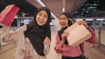 Two young Asian muslim friends feel happy showing shopping bags to camera, Modern Muslim lifestyle, capitalist consumer spending money, discounts sales products, enjoy shopping at the mall, city life