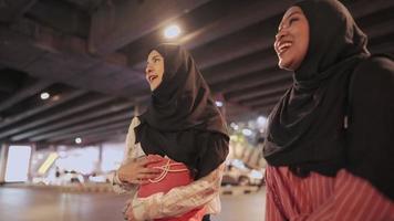 une jeune femme musulmane multiethnique parlant joyeusement en riant ensemble en attendant un taxi sur le bord de la route urbaine, traînant dehors avec un ami, ressemblance de filles, journée de shopping, porte des sacs en papier