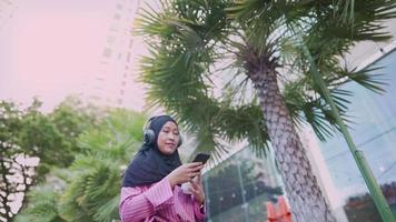 asiatisk attraktiv muslimsk kvinna bär hörlurar njut av att lyssna på musik medan du håller en kaffekopp som går längs vägkanten på gatan i moderna centrum under en dag, en bluetooth-anslutningsenhet, livet underhåller video