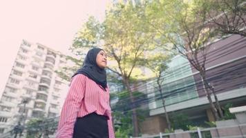 Asian female muslim wear black hijab stands on urban roadside in front of a modern glass office building with hand holding paper bags, people crossing a crosswalk, street traffic, green living space