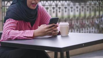 mulher muçulmana asiática usando telefone desfruta de momentos relaxantes na mesa de café, compartilhando histórias como postagem de comentários, sentado fora da zona do café, socializando em rede a cultura islâmica na sociedade moderna video