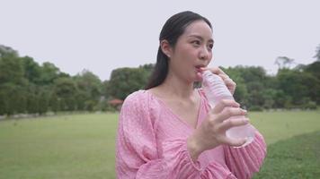 Young asian expectant mother drinking water uses straw sucking water from plastic bottle, late pregnancy stage, pregnant female drink water rehydration standing on the green grass inside open air park