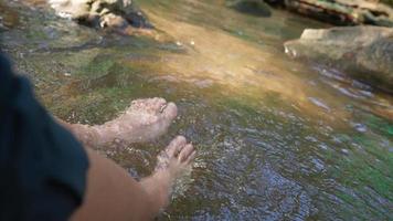 da vicino a piedi nudi ammollo nel fiume del canale della cascata, riposando durante le escursioni nel fine settimana, freschezza dalla natura, risorse naturali, gambe oscillanti per spruzzare l'acqua del ruscello, seduto sul bordo video