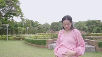 jolie femme enceinte asiatique porte une longue robe de mode rose embrassant caressant son gros ventre avec un visage souriant debout dans un parc naturel verdoyant seul, mère célibataire, anticipation du premier concept d'enfant video