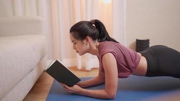 Aziatische fit meisje planking en leesboek multitasking, zelfmotivatie tijdens lockdown, thuis trainen oefening in de woonkamer met naast de bank, thuisonderwijs school bekijken studie lezing, warm licht