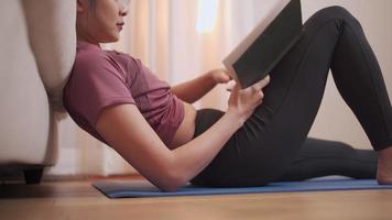 mujer asiática sentada leyendo un libro, después del ejercicio en casa, covid-19 encerrada, mujer sentada en una alfombra de ejercicio estudiando en la sala de estar con sofá, estudiante de educación en el hogar, actividad de ocio tiempo libre video