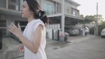 junge asiatische frau trägt ein weißes hemd, das durch die hausnachbarschaft läuft, vorbei an häusern und geparkten autos, spaß und freudige momente hat, den nachbarn die hände winkt, optimistische lebenspositive person video