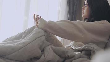 slaperige jonge aziatische vrouw zit gewoon op het bed in de slaapkamer in de ochtend, zijaanzicht, luie dag rommelig dekenbed witte gordijnen, natuurlijk daglicht zonnevlam lekt door het raam video