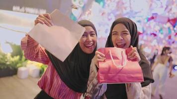 Two young asian excited Muslim women holding showing paper shopping bags while standing at shopping center inside modern downtown during the night, new lucky purchase bought on sale with discount video