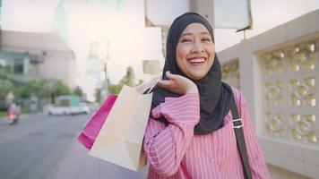 Young Asian Muslim wear black hijab holding shopping bags enjoy city life walking along street side pedestrian walk downtown, after work, modern Islam culture, smiling feel happy after spending money video