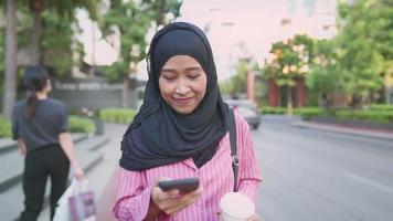 Young Asian Muslim wear using phone and holding coffee cup enjoy city life while walking along street side pedestrian walk downtown, after work, modern islam culture, Browsing the Web on her Device video