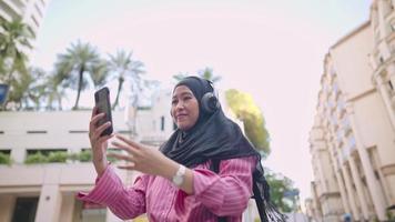 mulher muçulmana com lenço na cabeça usando telefone tomando selfie enquanto fica na beira da estrada no centro da cidade com um belo edifício alto único atrás, destino de viagem, um ambiente verde na capital video