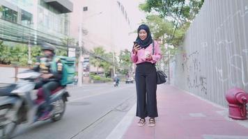 aantrekkelijke vrouwelijke aziatische moslim met afhaalmaaltijden koffiekopje en hoofd naar beneden scrollen op smartphone terwijl staande op een voetpad naast drukke straat, taxi-oproeptoepassing, gebruiker van gemakstechnologie