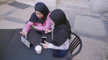 musulmanes asiáticos usan hiyab discutiendo el proyecto de trabajo en una tableta inalámbrica, siéntense para tener una conversación productiva en la mesa de café, trabajo en equipo en el espacio de los compañeros de trabajo, las niñas hablan a la hora del té de la tarde, ángulo alto video