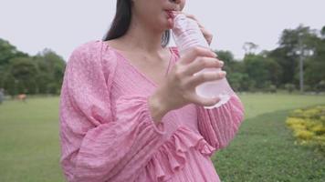 Eine fröhliche junge asiatische werdende Mutter, die Mineralwasser trinkt, während sie sich im grünen Park entspannt, einfaches Training für schwangere Frauen, späte Schwangerschaftsphase, sich darauf vorbereitet, Mutter zu sein, gesunder Lebensstil
