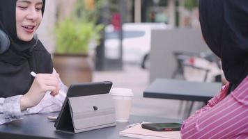 Young asian muslim friends wear hijab sitting outside having girls conversation in the afternoon tea break, casual daily lifestyle talking scene, using smart pen on touch screen, people and technology video