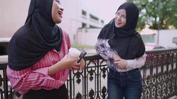 Two young asian Muslim women wear hijab having afternoon conversation standing on the road side and leaning on the fence friend meet up appointment, at coffee shop, friends talk islam culture society video