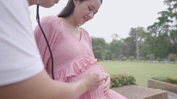 Closeup in beautiful moment of young parent using stethoscope listen to unborn baby inside mother's stomach, stroll at natural park, healthy breath in fresh air, waiting for new family member's coming video