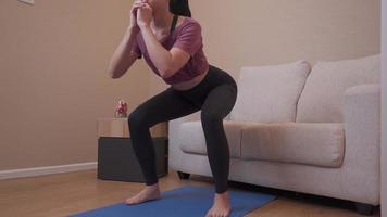 Female squatting on yoga mat in living room at home, standing exercise, stay home legs exercise, Healthy lifestyle during covid-19 quarantine, weight loss, body weight exercise, sofa on the background video