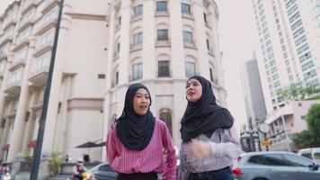 Two young Muslim girl friends walking together outside along downtown pedestrian walk, Islam ladies crossing street with traffic car vehicles on background, luxury city high-rise building, Arab wealth video