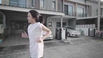 bela jovem asiática usa camisa branca correndo pelo bairro da casa passando por casas e carros estacionados, se divertindo e momentos alegres, condicionamento físico de motivação de fitness, exercício relaxante, video
