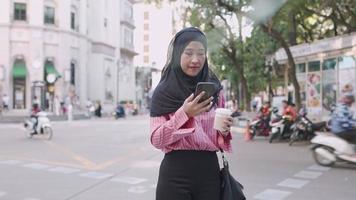 asiatische junge muslimische frau, die einen telefonanruf tätigt, kaffeetasse hält, die auf der straßenseite in der kaffeepause am nachmittag steht, tragbares mobiles gerät, geschäftige städtische szene in asien, taxi-abholservice anruft video