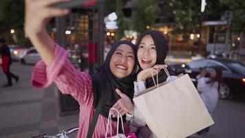 A young multi ethnic female Muslim joyfully taking selfie beside busy downtown road, with a view of skyscraper and city shops behind, non plastics bags concept, a friendship having fun memory concept video