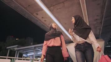 Two happy Southeast Asian muslim girls cheerfully walking together toward under a modern urban bridge, cultural unity friendship, happy and smiling while having positive conversation during weekend video