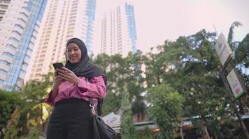 Aziatische vrouw in hoofddoek die telefoon gebruikt om vrolijk naar een vriend te sms'en terwijl ze langs de weg in het centrum van de stad staat, hoogbouw, moderne schone stedelijke architectuur, groene omgeving in de hoofdstad video