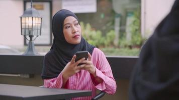 los jóvenes musulmanes asiáticos se sientan en la mesa de café al aire libre usando un teléfono móvil, reciben mensajes de texto en línea, las redes sociales leen cosas en línea, usan ropa tradicional islámica hiyab, musulmanes modernos