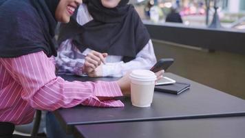 zwei junge asiatische muslimische mädchen, die sich tagsüber am cafétisch unterhalten, fröhliche attraktive frauen mit schwarzem hijab lächeln und lachen zusammen, hand mit touchscreen-telefon, moderner junger lebensstil video