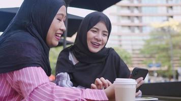 Young asian islam friends wear hijab sitting at outdoor cafe on their afternoon coffee break showing new smartphone, online shopping application, friends conversation gossip, people and technology video