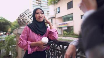 mulher muçulmana asiática gosta de conversar com amigos, comunidade islâmica, segurando a xícara de café, fazendo uma pausa no centro da cidade, café ao ar livre, tráfego atrás mostrando o fundo da rua urbana, sorrindo e divertido video