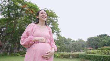 giovane madre asiatica felice che cammina nel parco all'aperto, strofinandosi parlando con il suo primo figlio all'interno della grande pancia incinta, 38 settimane di gravidanza ultima fase prima del parto, calore familiare legame d'amore