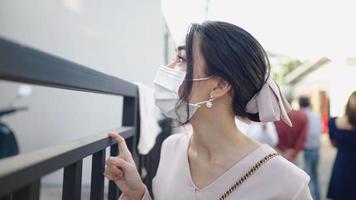 Portrait of asian beautiful girl waiting in front of metal house fence, a well dressed woman with face mask stand isolated from crowded behind turning self and looking with smiling eyes to came video