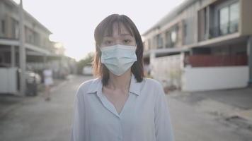 Asian woman wear face mask walking in front of house neighborhood street, Corona virus covid19 awareness caution, standing outdoor sunset social distancing concept, showing confidence by thumbs up