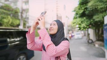 Exited asian muslim female wear black headscarf stands taking a modern cityscape photo on busy roadside during a sunny day, asia travel and memories, people and convenience wireless technology video