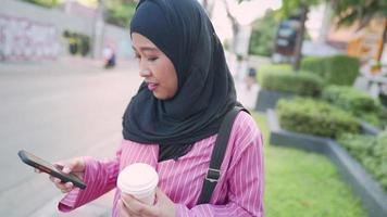 Young Asian Muslim wear hijab phone and holding coffee cup, city life standing on street side pedestrian walk downtown, after work, modern Islam culture, use navigation application, online shopping video