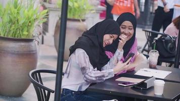 duas mulheres muçulmanas asiáticas usam hijab na pausa para o café da tarde usando um novo smartphone, compartilhando conhecimento, sentados fora da zona externa do café, redes sociais, postando fotos online video