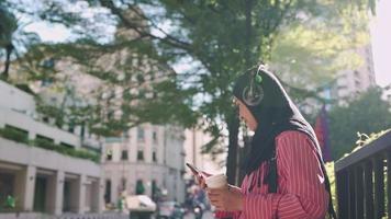 asiatische muslimische frau trägt kopfhörer, hört podcast, warmer sonniger tag, hält kaffeetasse auf der straßenseite nachmittagskaffeepause, benutzt smartphone, genießt musik und outdoor-vibe