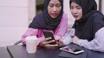 twee aziatische moslims dragen hijab vrije tijd zittend aan de cafétafel, pronken met nieuwe mobiele telefoon, aanraken op smartphone-tocuh-scherm, draadloze draagbare technologie-apparaten in de moderne samenleving video