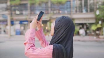 Rear view of a black hijab muslim woman stands on urban street roadside holding phone taking photo of a city modern architecture, distant communication, online internet connection, travel and memories