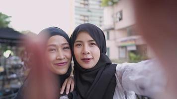 Young asian Muslim girls friend wear hijab holding phone FPV shot taking selfies enjoying travel trip, standing on the street side feeling happy and fun, phone front camera self portrait video