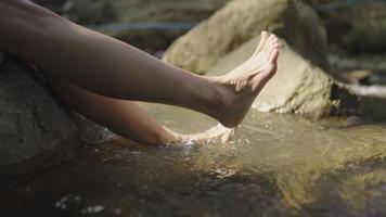 giovane donna seduta sul bordo delle rocce inzuppando le gambe nell'acqua della cascata del ruscello, godendosi con acqua fresca e fresca nel torrente, spruzzando acqua fresca pulita e soleggiata, fuga dalla città video